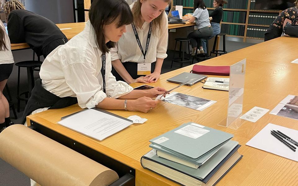 Twee kunstgeschiedenisstudenten analyseren samen een afbeelding aan een tafel vol boeken, prints en onderzoeksdocumenten op het rkd -  nederlands instituut voor kunstgeschiedenis.