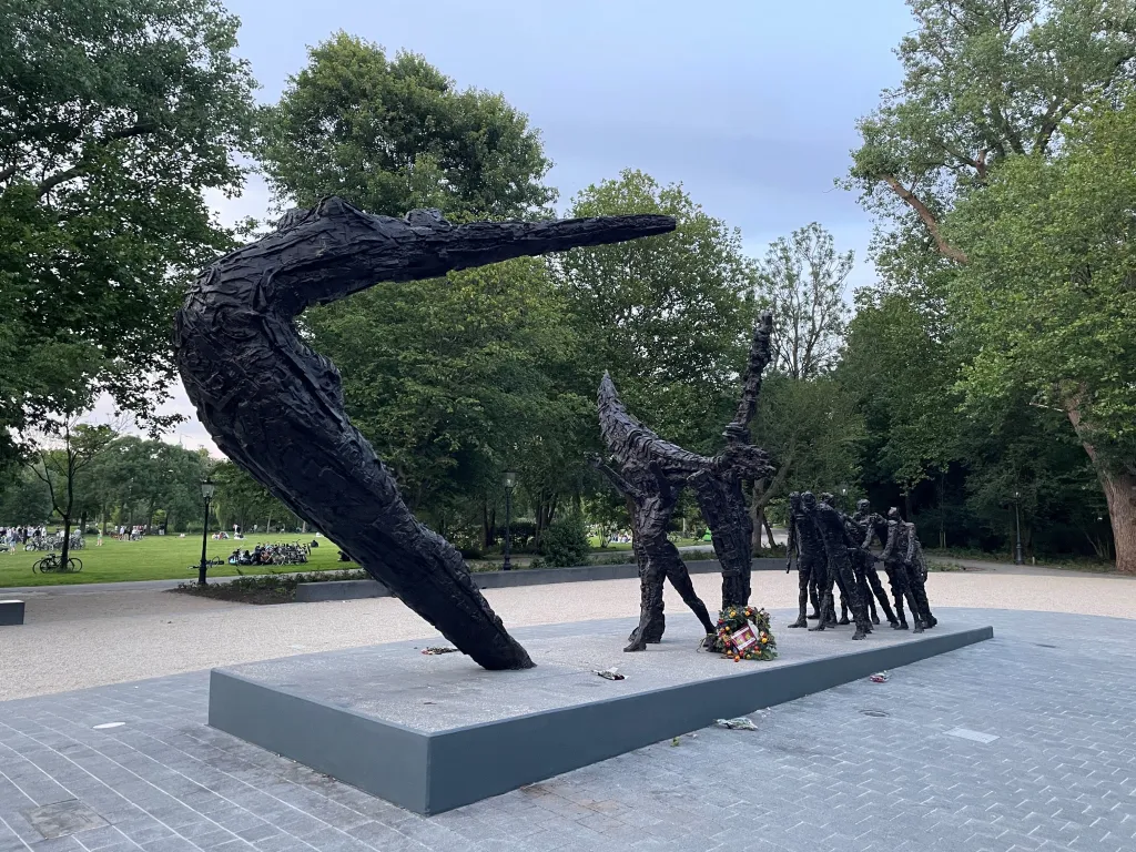 Nationaal Monument Slavernijverleden. Beeld van een vrouwfiguur met haar hoofd naar boven en armen gestrekt in de lucht. Daarachter meer figuren die kleiner zijn. Op de achtergrond een park met bomen.