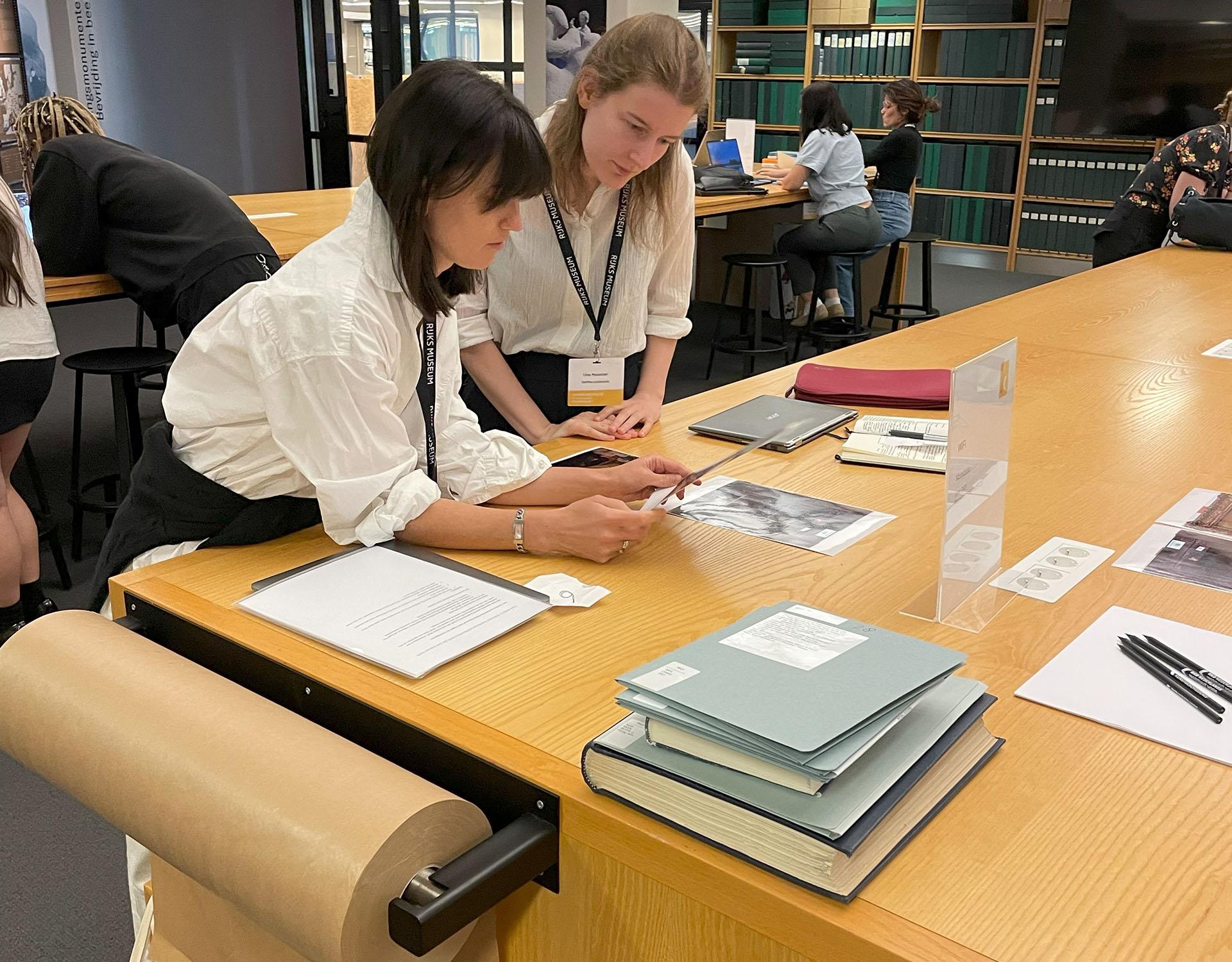 Twee kunstgeschiedenisstudenten analyseren samen een afbeelding aan een tafel vol boeken, prints en onderzoeksdocumenten op het rkd -  nederlands instituut voor kunstgeschiedenis.